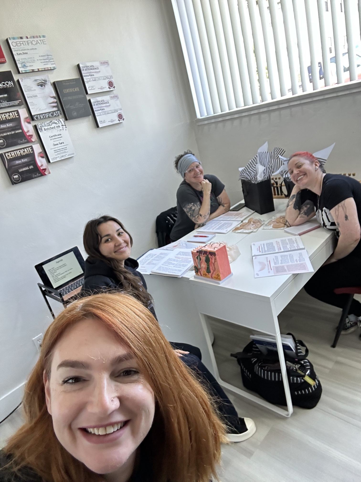Anna taking a selfie in the downtown classroom with three students at a white table with course binders and a skin model