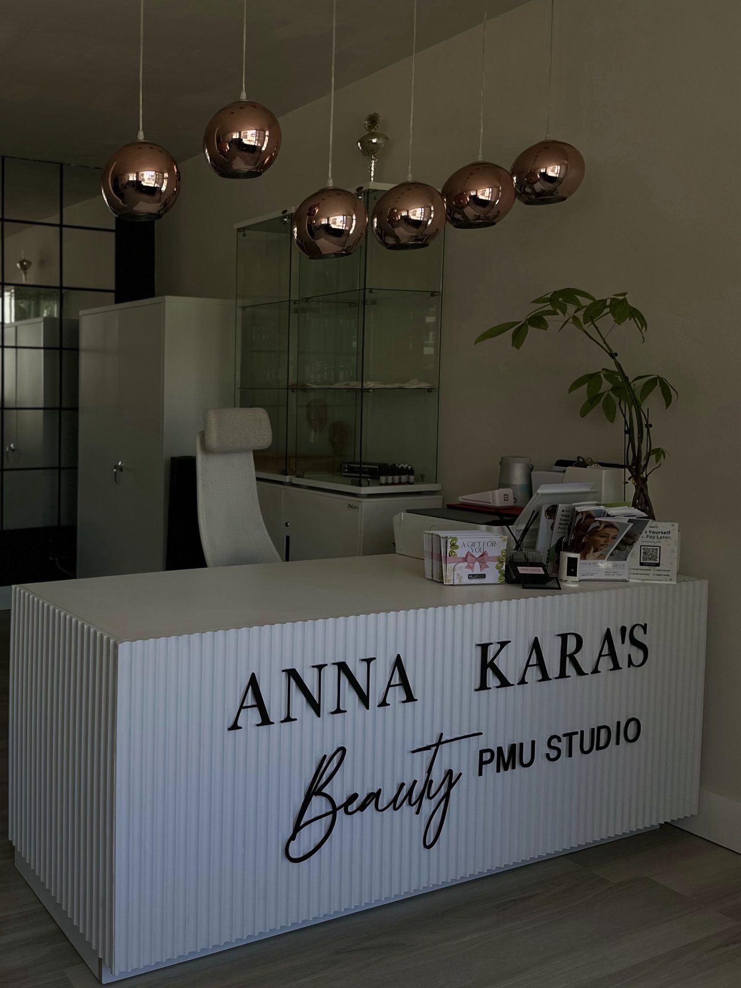 Reception desk reading Anna Kara's Beauty PMU Studio under copper pendant lamps