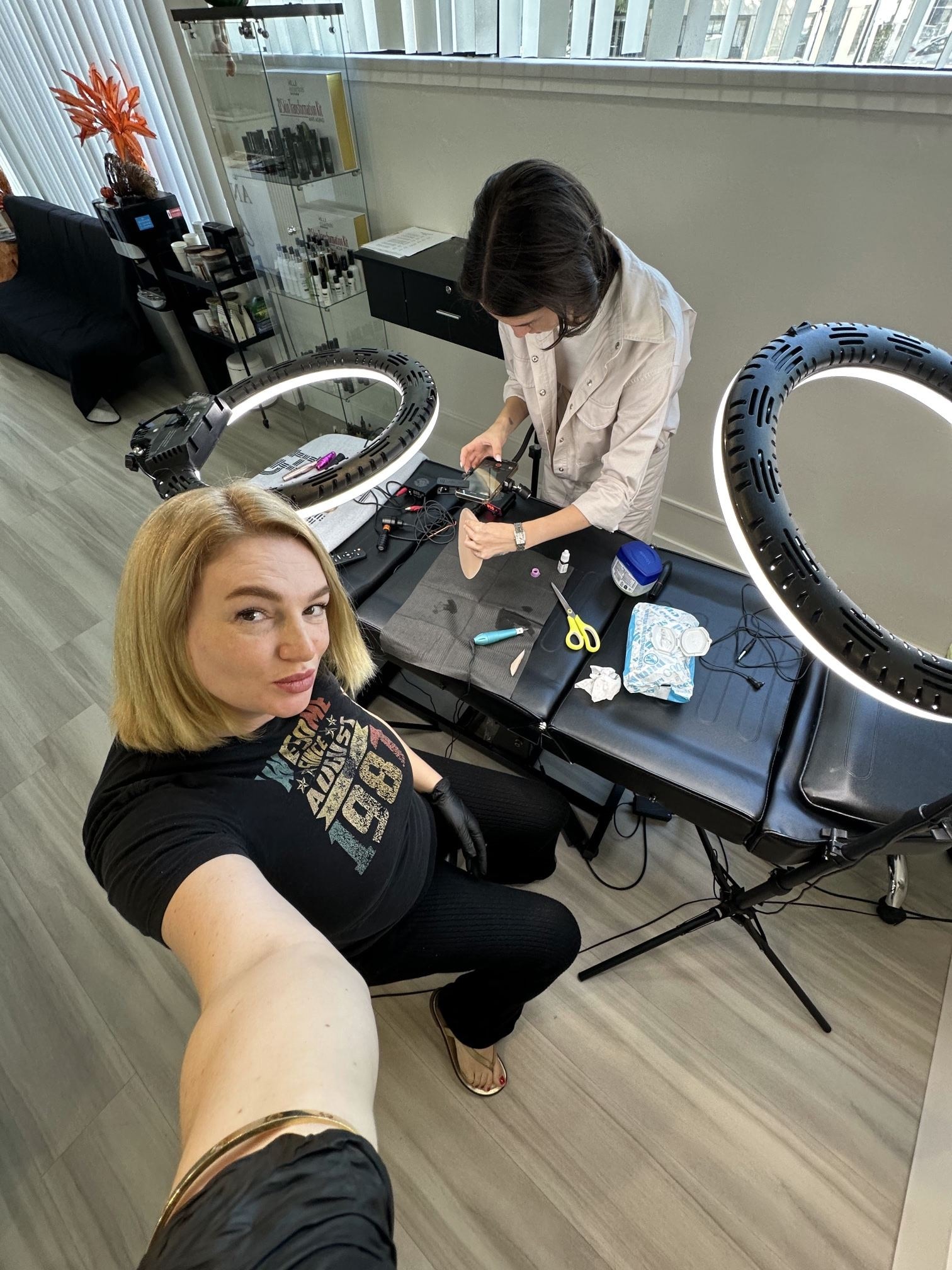 Anna in a black T-shirt in the foreground while a student works on a practice skin pad under ring lights during a paramedical class