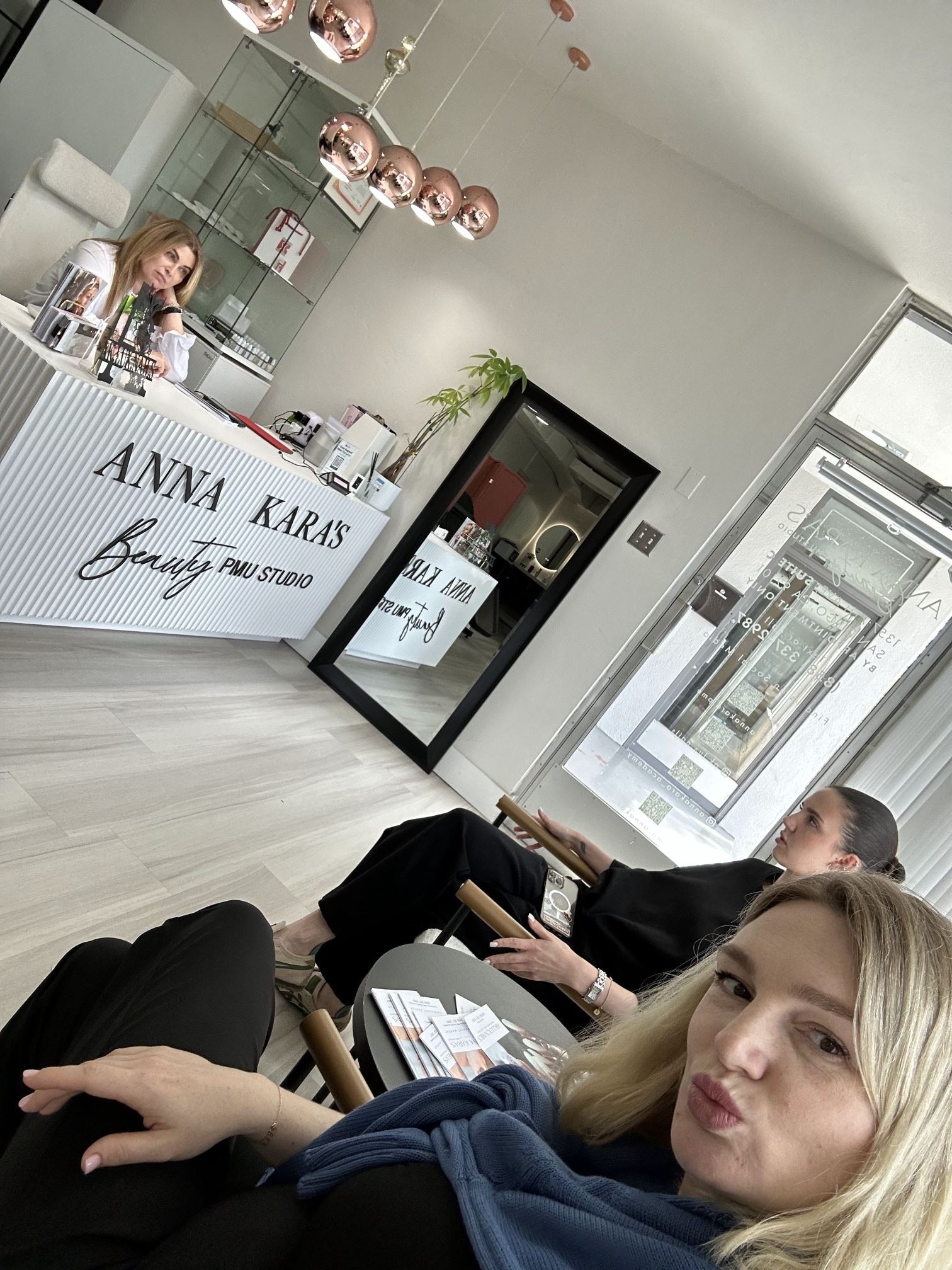 Mila, the administrator, at the white ribbed reception desk of Anna Kara's Beauty PMU Studio, with Anna and a team member in the waiting area