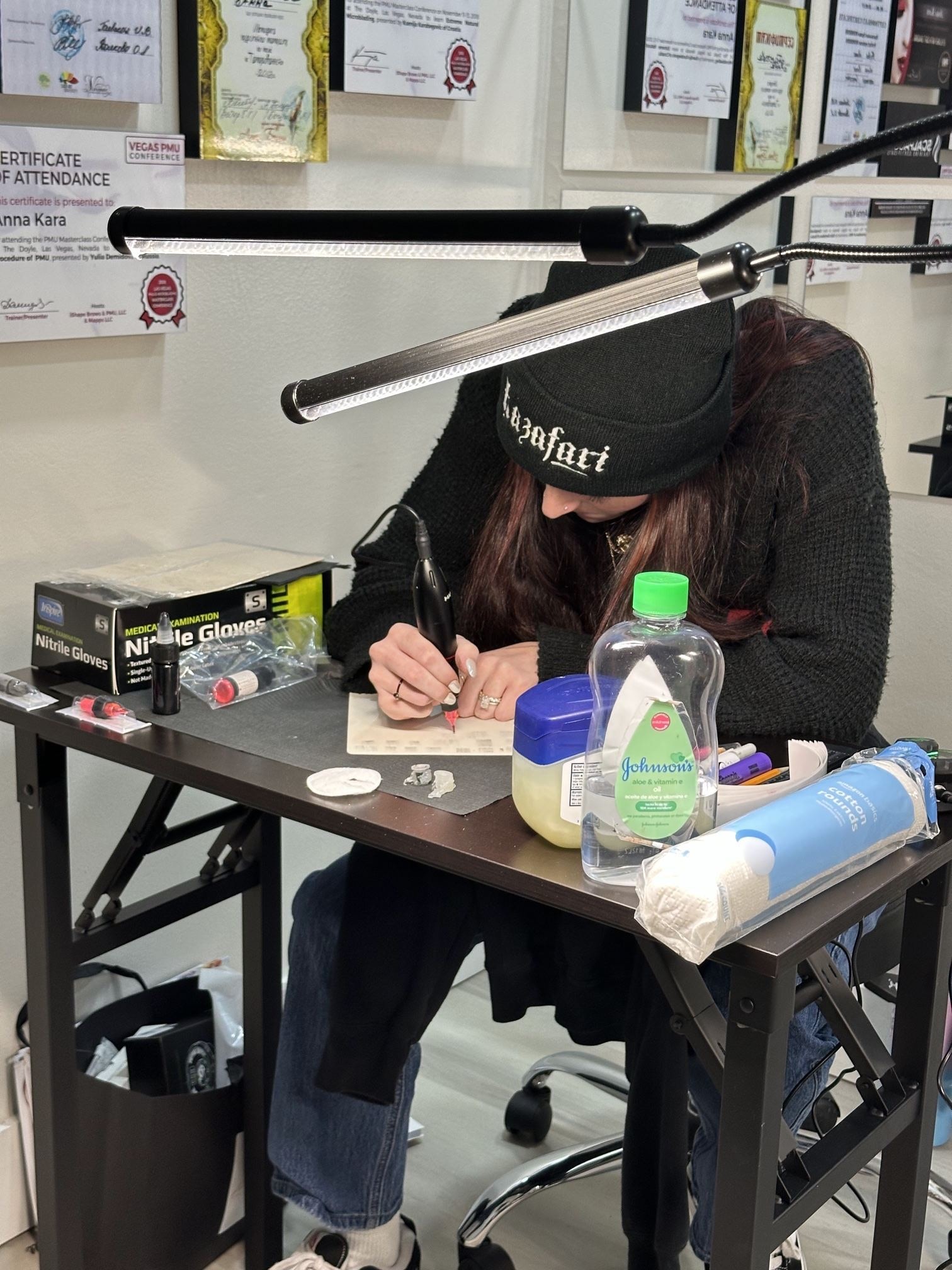 A student in a black beanie bends over a desk under a lamp, practicing with a pigment machine on a latex practice skin